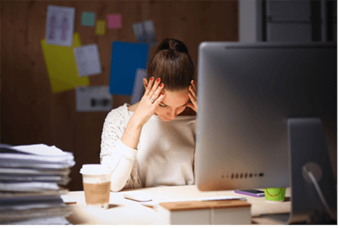 Woman sitting on a desk in front of a computer with a stack of papers feeling stressed out